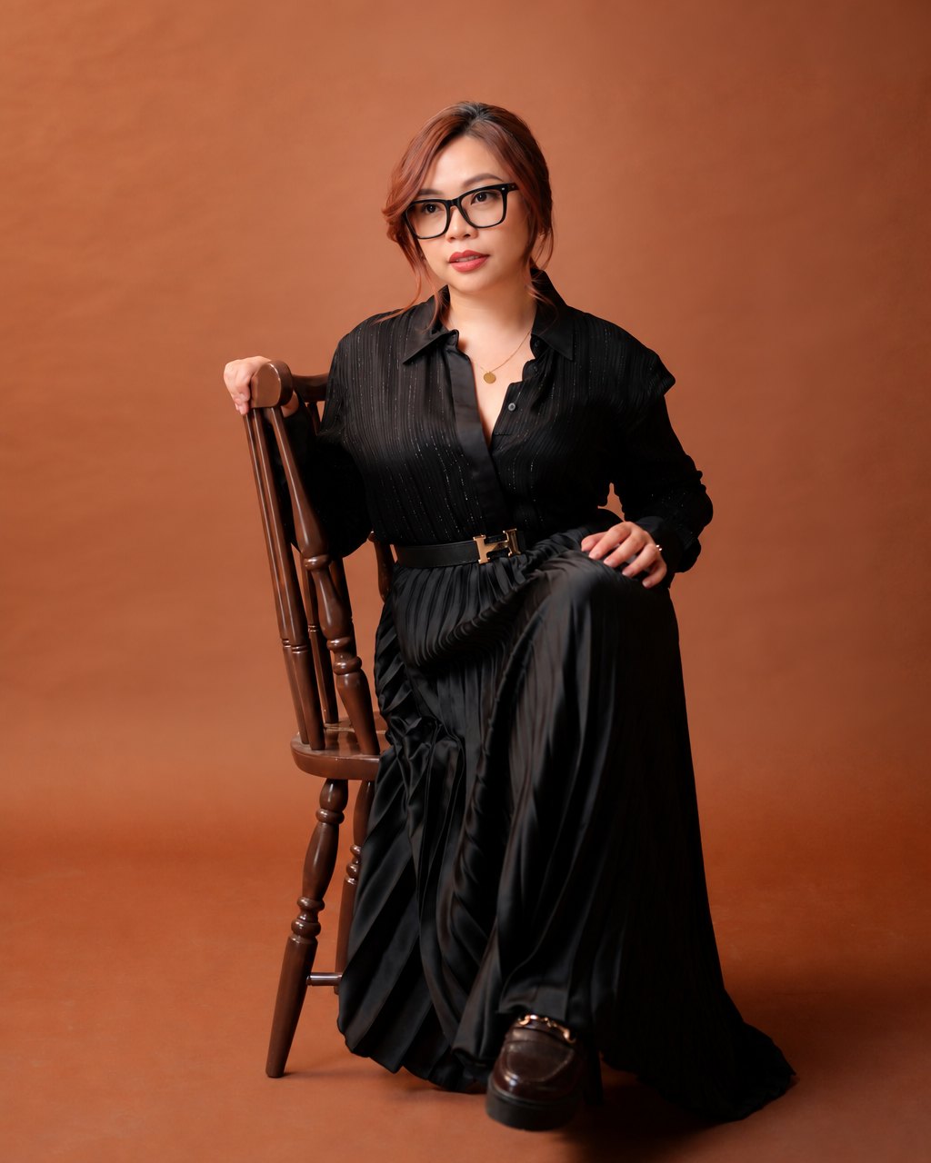 Nikki "Qian Xi" Jurado-Mancilla in glasses and an all-black outfit, seated on a wooden chair against a warm brown studio backdrop